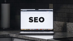 Laptop displaying large black "SEO" text on a white document, positioned on a concrete desk in a modern workspace.