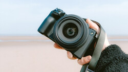 Photographer holding a mirrorless camera with attached lens against a beach backdrop.