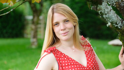 Young woman with blonde hair wearing a red polka dot dress in an outdoor garden setting.
