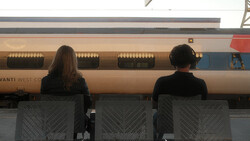 Two people seated facing a parked train with tan and blue livery, shot from behind through mesh chairs.