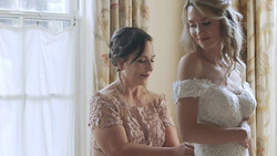 Two women in elegant lace dresses share a moment by a bright window with floral curtains.