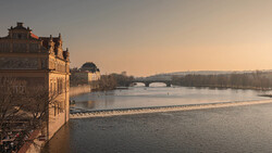 Ornate riverside building at golden hour overlooking a calm river with an arched bridge in the distance.