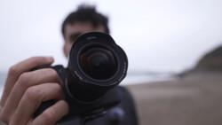 Photographer holding a camera with telephoto lens pointed toward the camera in a beach setting.