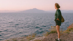 Runner in green jacket standing on coastal cliff at sunset, facing distant snow-capped volcano across water.