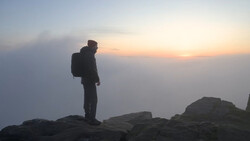 Silhouetted hiker standing on rocky outcrop against golden sunset sky with atmospheric haze.