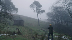 Photographer in yellow beanie shooting in misty hillside landscape with solitary pine tree and stone structure.