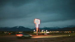 Neon-lit roadside sign glowing against dark mountains at dusk with car light trails.