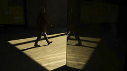 Person walking through geometric patches of golden sunlight in a minimalist architectural space.