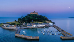 Coastal castle on a forested island surrounded by a harbor with moored boats at twilight.