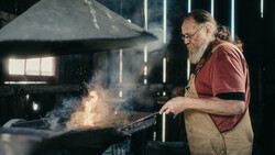 Blacksmith working at an anvil with sparks flying in a workshop lit by overhead lighting.