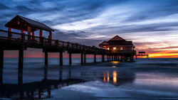 Illuminated pier structures silhouetted against a vibrant sunset sky reflecting in shallow water.