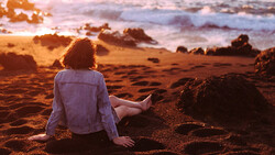 Person sitting on beach sand facing ocean at golden hour sunset.