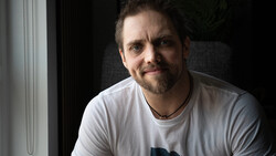 Man with goatee wearing light gray t-shirt, photographed against dark background with window light.
