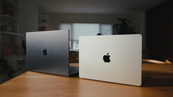 Two MacBook laptops displayed closed on a wooden desk, one silver and one white, photographed in indoor studio lighting.