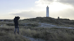 Photographer with camera raised to face standing in coastal dunes with lighthouse on distant hill.