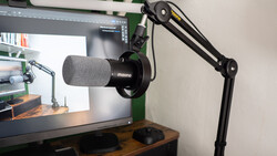 Boom arm microphone setup mounted above wooden desk with monitor displaying editing software.