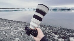 Photographer holding a telephoto lens with black and white striped sections over an icy glacial lagoon.
