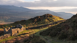 Golden-hour sunlight illuminates a rocky hilltop with ancient stone ruins overlooking a verdant valley and distant mountains.
