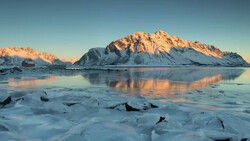 Snow-covered mountain reflected in partially frozen water at golden hour.
