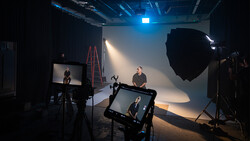Professional film production studio setup with subject seated in key light, multiple cameras and monitors monitoring the shoot.