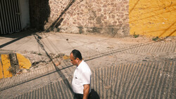 Man in white shirt standing in sunlit alley with geometric shadows and yellow wall.