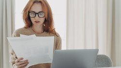 Woman with reddish-brown hair and glasses reviewing documents while working at a laptop.