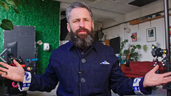 Bearded man in dark denim jacket gesturing expressively in a creative studio space.
