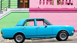 Bright blue vintage sedan parked against contrasting green and pink building facade.