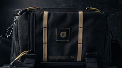 Black camera bag with tan stripes and brass hardware against dark background.
