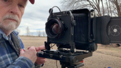 Photographer holding a large format camera with bellows and ground glass in an open field.