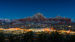 Snow-dusted red rock formations rise above a town illuminated by warm lights at dusk.