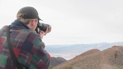 Photographer with camera raised to face, wearing plaid shirt and cap, standing on hillside overlooking desert valley.