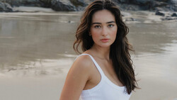 Young woman in white tank top posing at beach with rocky outcrops behind her.