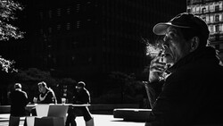 Man in baseball cap smoking cigarette in urban plaza with other figures blurred in background.