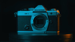 OM System OM-3 rangefinder camera with cyan and orange gel lighting against dark background.