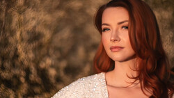 Woman with reddish-brown hair against a textured brown background, wearing a light-colored knit sweater.