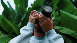 Photographer holding a telephoto lens up to frame a shot among large green leaves.