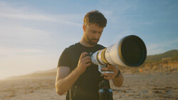 Photographer holding a telephoto lens on a beach during golden hour.