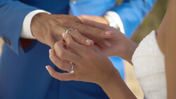 Bride and groom exchanging wedding rings, hands clasped together with blue suit and white dress visible.