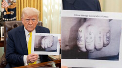 Man in business suit holding up photographs of tattooed knuckles with letters.
