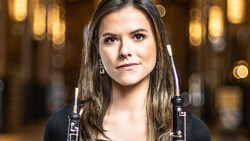 Portrait of a young woman holding oboes against a warm, blurred interior background.