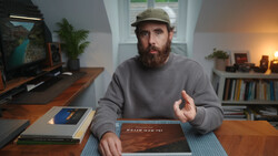 Bearded man wearing a gray sweatshirt and cap gestures while discussing photography books at a wooden desk.