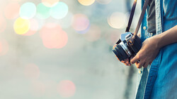 Photographer in denim shirt holding a vintage rangefinder camera with soft-focus bokeh lights in background.
