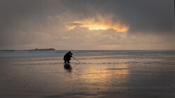 Silhouetted figure wading in shallow water on a beach at sunset, with dramatic golden clouds breaking through overcast sky.