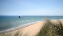 Sandy beach with dune grasses in foreground, calm turquoise water, and distant lighthouse on the horizon.