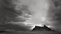 Silhouetted rocky pinnacles rise from a flat desert plain beneath a dramatic storm sky.