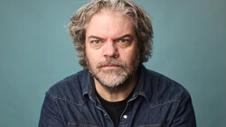 Man with curly gray hair and beard wearing a denim shirt against a teal background.