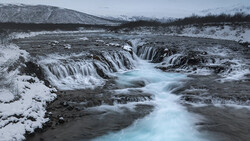Frozen waterfall cascading through snow-covered landscape with turquoise glacial water flowing between dark rocks.