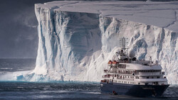 Research vessel positioned before a massive ice shelf with towering white cliffs and deep crevasses.