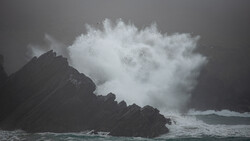 Massive ocean wave crashes against dark rocky coastline, creating explosive white spray and mist.